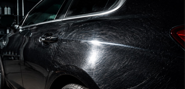 A close-up of a glossy black car body panel showing fine scratch marks and swirl marks on the paint, with a dark garage background.